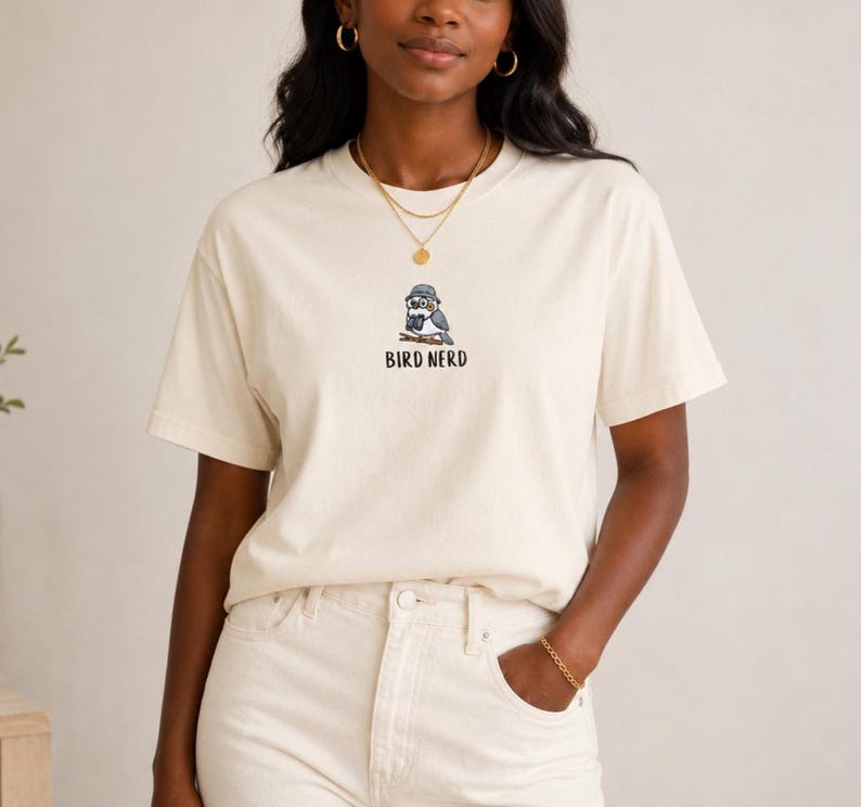 May include: Cream-colored t-shirt featuring a cartoon bird wearing glasses and a hat, perched on a branch. The text "BIRD NERD" is printed below the bird illustration in black. The shirt is made of a soft material.