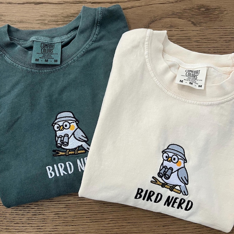 May include: Two folded t-shirts, one teal and one cream-colored, featuring an embroidered bird design with the text "BIRD NERD". The bird wears glasses and a hat, holding binoculars. Both shirts have a "Comfort Colors" tag.