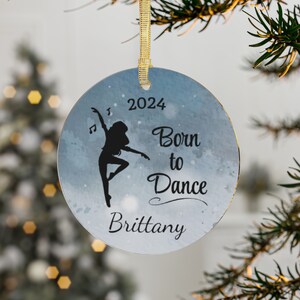 May include: A round, blue and white Christmas ornament with a silhouette of a dancer and the text "Born to Dance" and "2024". The name "Brittany" is written at the bottom of the ornament.
