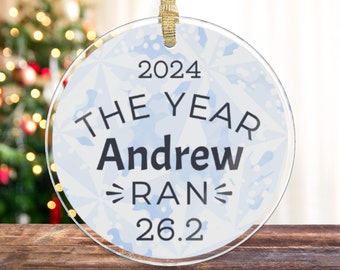 May include: A clear round glass ornament with a blue and white snowflake design. The ornament has the text "2024 The Year Andrew Ran 26.2" printed on it.