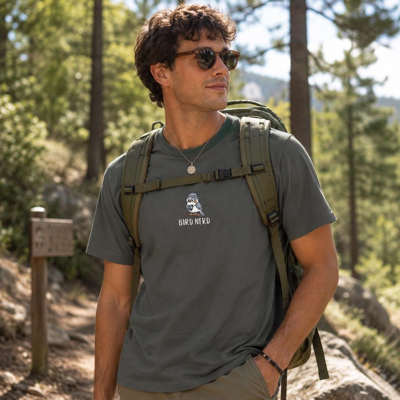 May include: Dark green t-shirt with a cartoon bird wearing a hat and the text "BIRD NERD". The shirt is made of a soft material and is being worn outdoors.
