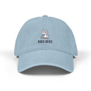 May include: Light blue baseball cap with a curved brim. The cap features an embroidered design of a bird wearing glasses and a hat, holding a twig, with the text "BIRD NERD" below the bird.