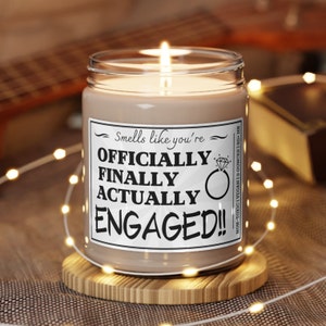 May include: A brown candle with a white label that reads "Smells like you're OFFICIALLY FINALLY ACTUALLY ENGAGED!!" The candle is lit and sitting on a wooden coaster.