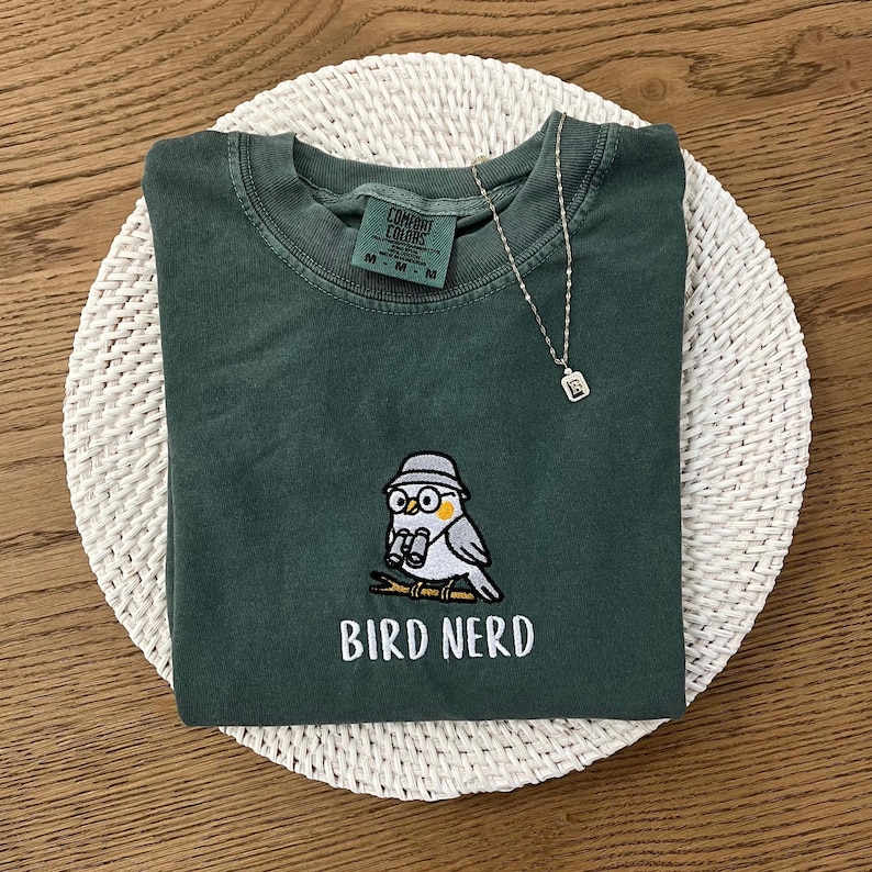 May include: A dark green t-shirt with a cartoon bird design and the text "BIRD NERD" in white. The bird is wearing a hat and glasses. A silver necklace with a small pendant is also present.