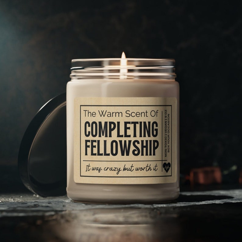 Puede incluir: Una vela marr&oacute;n con una etiqueta blanca que dice "The Warm Scent Of Completing Fellowship. It was crazy but worth it." La vela est&aacute; en un tarro de vidrio transparente con una tapa negra.