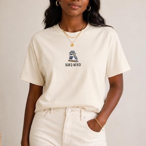 May include: Cream-colored t-shirt featuring a cartoon bird wearing glasses and a hat, with the text "BIRD NERD" below. The shirt is made of a soft material and has short sleeves. The model is wearing light-colored pants.