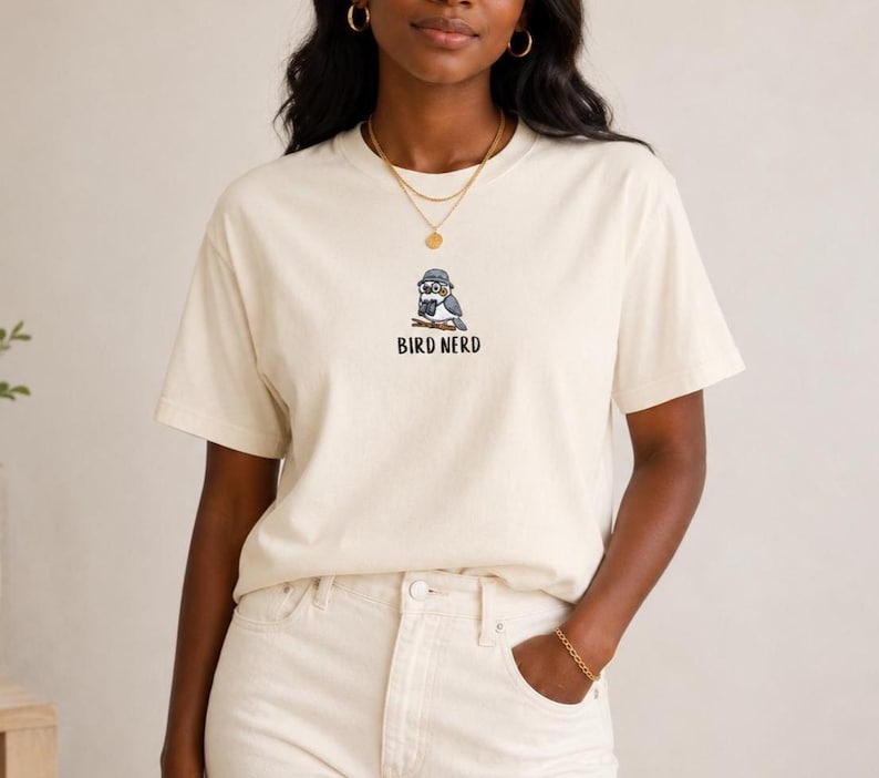 May include: Cream-colored t-shirt featuring a cartoon bird wearing glasses and a hat, with the text "BIRD NERD" below. The shirt is made of a soft material and has short sleeves. The model is wearing light-colored pants.