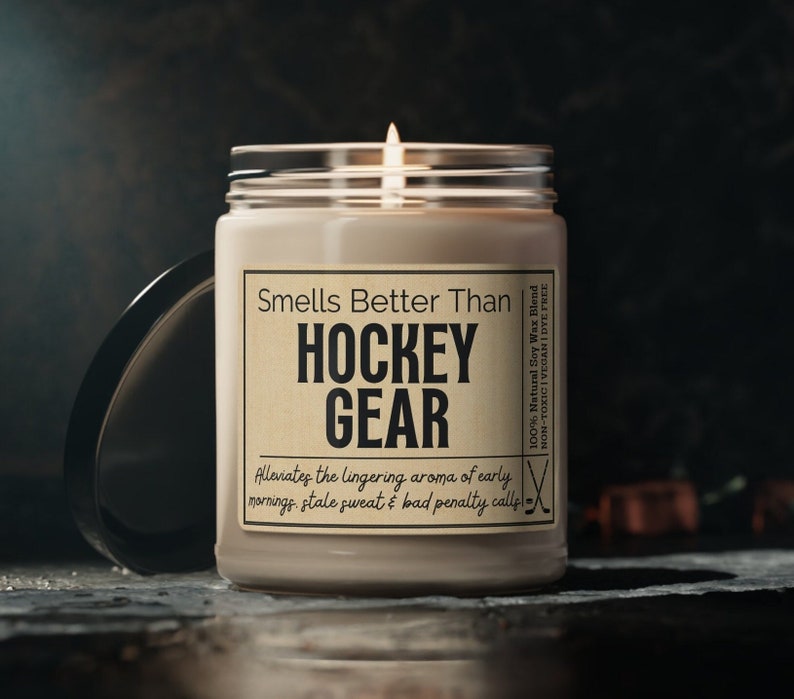 May include: A brown candle with a white label that reads "Smells Better Than HOCKEY GEAR Alleviates the lingering aroma of early mornings, stale sweat & bad penalty calls." The label also includes the text "100% Natural Soy Wax Blend NON-TOXIC | VEGAN | DYE FREE" and a hockey stick graphic.