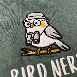 May include: Embroidered patch featuring a cartoon bird wearing glasses and a hat, holding binoculars, perched on a branch. The bird is gray, white, and yellow, with the text "BIRD NERD" below in white.