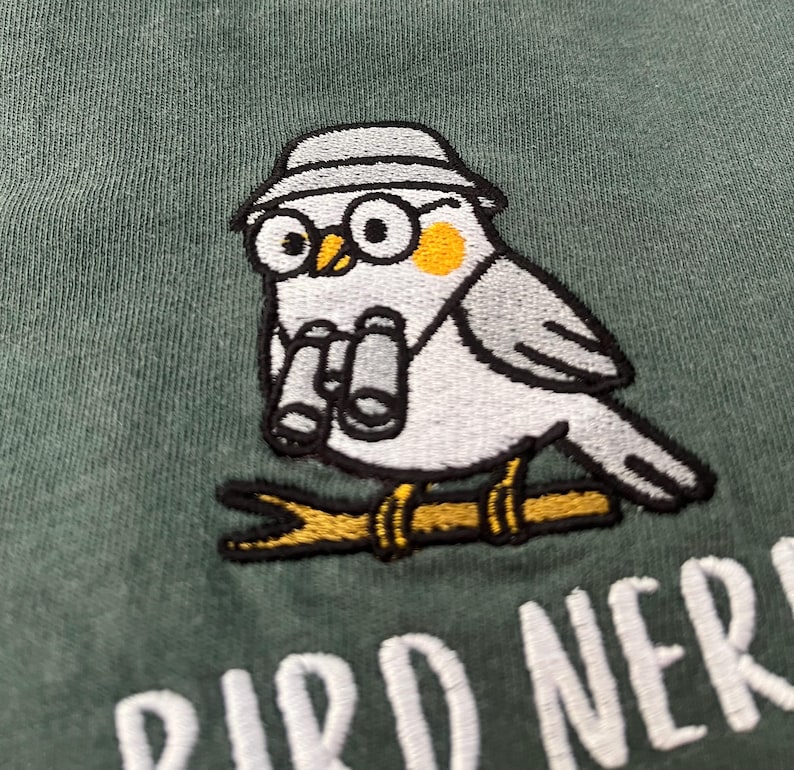 May include: Embroidered patch featuring a cartoon bird wearing glasses and a hat, holding binoculars, perched on a branch. The bird is gray, white, and yellow, with the text "BIRD NERD" below in white.