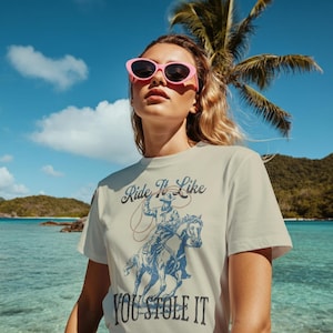 Peut inclure: T-shirt crème avec un motif bleu d'un cow-boy à cheval et le texte "Ride It Like You Stole It". La personne porte des lunettes de soleil roses, avec une plage tropicale en arrière-plan.