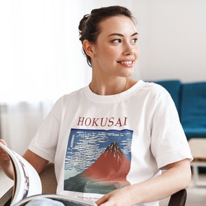May include: A woman wearing a white t-shirt with a graphic print of a mountain and the text "HOKUSAI". She is sitting in a chair and holding a magazine.