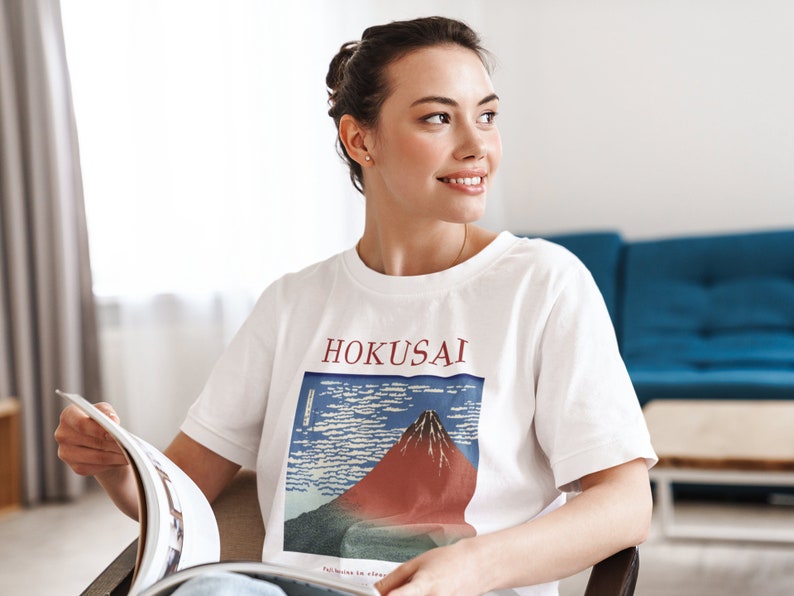 May include: A woman wearing a white t-shirt with a graphic print of a mountain and the text "HOKUSAI". She is sitting in a chair and holding a magazine.