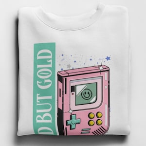 May include: White sweatshirt with a green rectangle that says "Old But Gold" and a pink and green retro handheld game console with a smiley face on the screen.