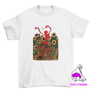 以下が含まれることがあります： 黄色のひまわり畑で踊る赤い骸骨が描かれた白いTシャツ。骸骨は赤色で骨は白く、三日月を手に持っています。