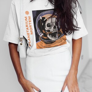 May include: White t-shirt with a graphic print of a skull in a space helmet. The text "PLANETARIUM MORTEM" is printed vertically on the left side of the graphic.
