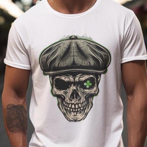 Puede incluir: Una camiseta blanca con un estampado gráfico de una calavera con una gorra plana. La calavera tiene un ojo con un trébol de cuatro hojas verde.