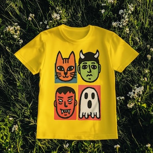 May include: A yellow t-shirt featuring four cartoon faces: an orange cat, a green devil, a red monster, and a white ghost. The faces are in individual colored squares. The shirt is displayed on a bed of green grass.