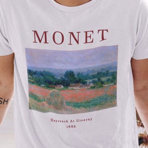 May include: White t-shirt with a painting of a landscape by Claude Monet. The painting is titled "Haystack At Giverny 1886".