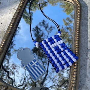 Peut inclure: Un collier et un porte-clés assortis avec un motif à carreaux bleu et blanc. Le collier est en forme de petite pochette, et le porte-clés a un motif similaire. Les deux sont présentés sur un miroir doré.