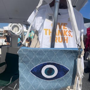 May include: A light blue denim clutch bag featuring a large, beaded evil eye motif. The bag has a zip closure and a white tassel. A white t-shirt with the words "GIVE THANKS, RUH" is visible in the background.