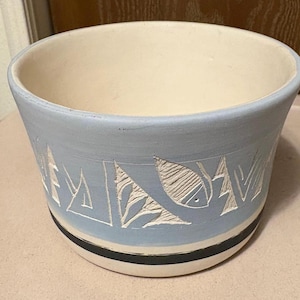 May include: A light blue ceramic planter with a cream-colored interior. The exterior features carved white geometric and fish designs. A black and white stripe accents the base.