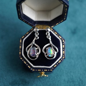 Könnte beinhalten: Ein Paar silberne Ohrhänger mit schillernden Opal-Steinen, die in einem filigranen, gedrehten Design gefasst sind. Die Ohrringe werden in einer blauen Samt-Schmuckschatulle präsentiert.