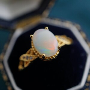 May include: A gold ring featuring a large oval opal gemstone set in a delicate, ornate design. The ring is displayed in a black velvet ring box.