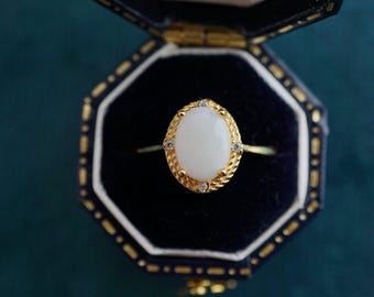 May include: A gold ring with a white oval gemstone surrounded by smaller clear gemstones. The ring is in a black velvet ring box.