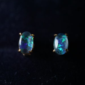 May include: A pair of oval opal stud earrings. The opals show a vibrant play of colour with flashes of blue and green. The earrings are set in gold-coloured prongs against a dark background.