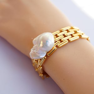 Op de afbeelding: Een gouden armband met een grote, onregelmatige parel. De armband is gemaakt van een reeks met elkaar verbonden gouden staven.