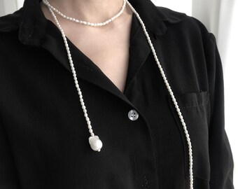 May include: A long pearl necklace with two strands, one shorter than the other, worn on a black shirt. The necklace has a simple design and is made of white pearls.