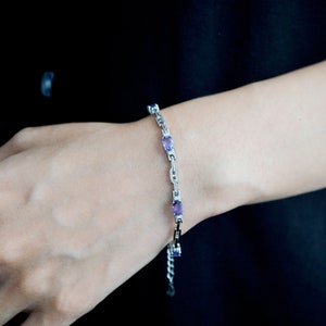 May include: A silver bracelet with oval purple gemstones. The bracelet has a delicate chain design with the gemstones evenly spaced. The bracelet is displayed against a dark background.