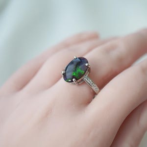 May include: A silver ring with a large, oval black opal displaying green and blue hues. The band is set with small, sparkling stones. The ring is shown on a hand, highlighting the jewellery's design and colour.