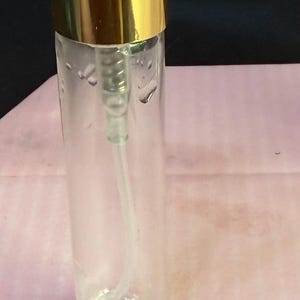 Op de afbeelding: Een heldere glazen monsterfles met een goudkleurige dop en sproeier. De fles heeft een label met de tekst "SAMPLE 5ml". De fles staat op een roze ondergrond.
