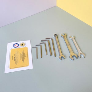 May include: A set of seven tools, including wrenches and Allen keys, laid out on a light blue surface. The tools are silver and have a shiny finish.