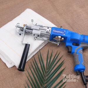 May include: A blue and grey electric tufting gun with a black handle and a metal needle mechanism. The device features a label reading TuftingGuide and is positioned next to a folded white fabric with thin orange lines and a green palm leaf.