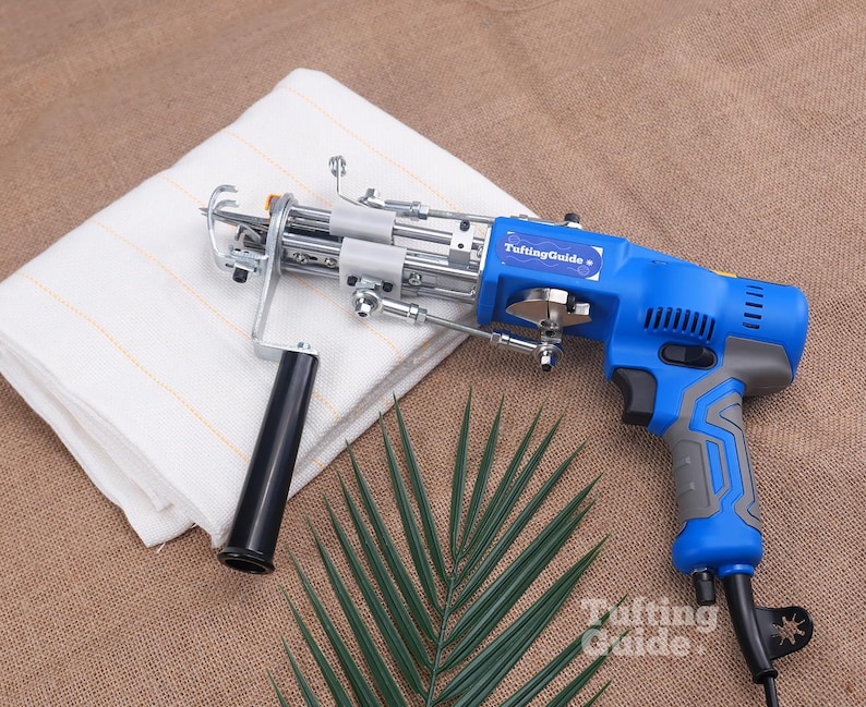 May include: A blue and grey electric tufting gun with a black handle and a metal needle mechanism. The device features a label reading TuftingGuide and is positioned next to a folded white fabric with thin orange lines and a green palm leaf.