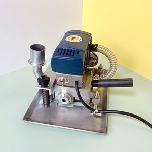 May include: A blue and silver electric edge banding machine with a black handle and a clear plastic hose. The machine has a metal base and a metal exhaust pipe.
