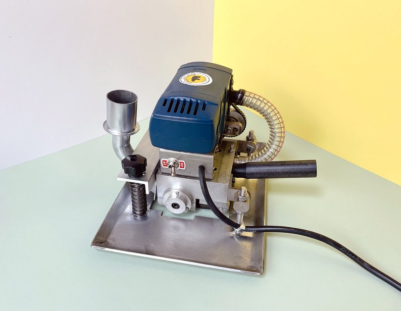 May include: A blue and silver electric edge banding machine with a black handle and a clear plastic hose. The machine has a metal base and a metal exhaust pipe.