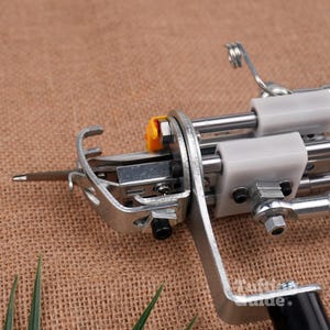 May include: A metal tufting machine component featuring a silver needle, a curved metal guide, and white plastic blocks mounted on parallel steel rods. A small yellow plastic piece is attached near the needle mechanism, and a black handle is visible.