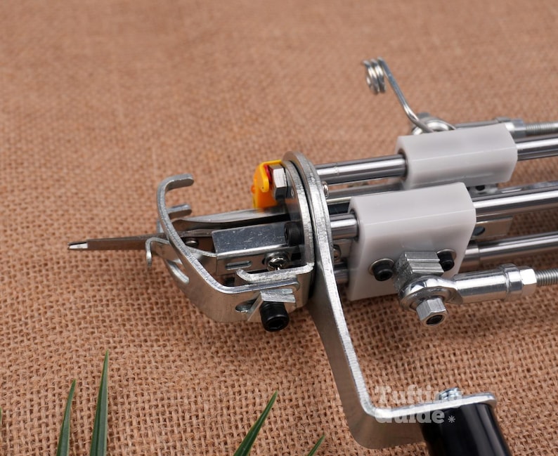 May include: A metal tufting machine component featuring a silver needle, a curved metal guide, and white plastic blocks mounted on parallel steel rods. A small yellow plastic piece is attached near the needle mechanism, and a black handle is visible.