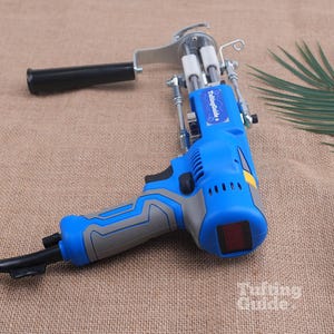 May include: A blue and grey electric tufting gun with a black handle, a metal needle mechanism, and a power cord. The device features a blue label with the white text TuftingGuide and a small red display screen on the back of the motor housing.