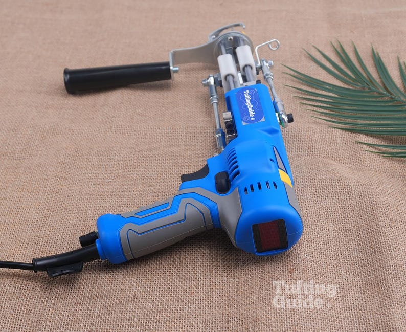 May include: A blue and grey electric tufting gun with a black handle, a metal needle mechanism, and a power cord. The device features a blue label with the white text TuftingGuide and a small red display screen on the back of the motor housing.