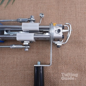 May include: A metal tufting machine featuring a blue handle, silver mechanical components, white plastic guides, and a black grip. The device includes a needle mechanism with a metal guard and visible screws. The text Tufting Guide is visible.