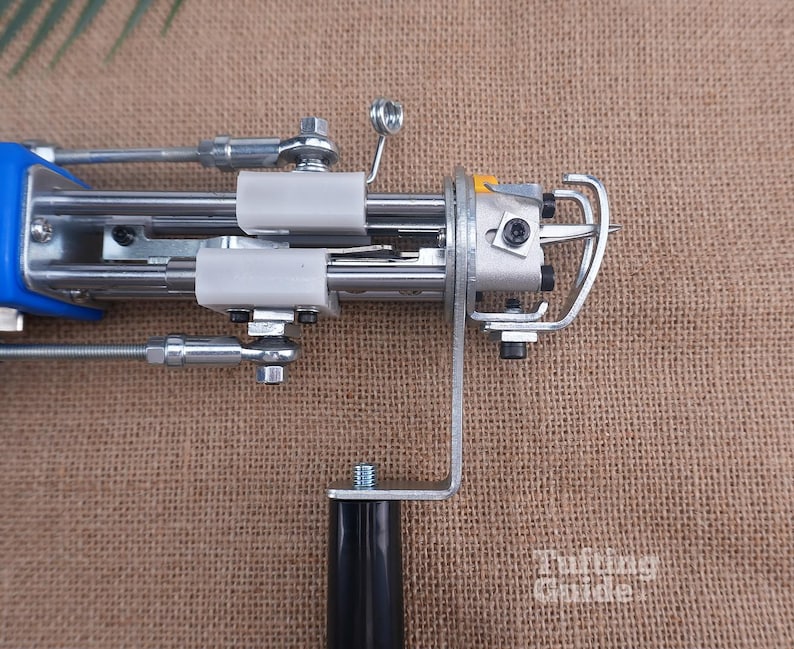 May include: A metal tufting machine featuring a blue handle, silver mechanical components, white plastic guides, and a black grip. The device includes a needle mechanism with a metal guard and visible screws. The text Tufting Guide is visible.
