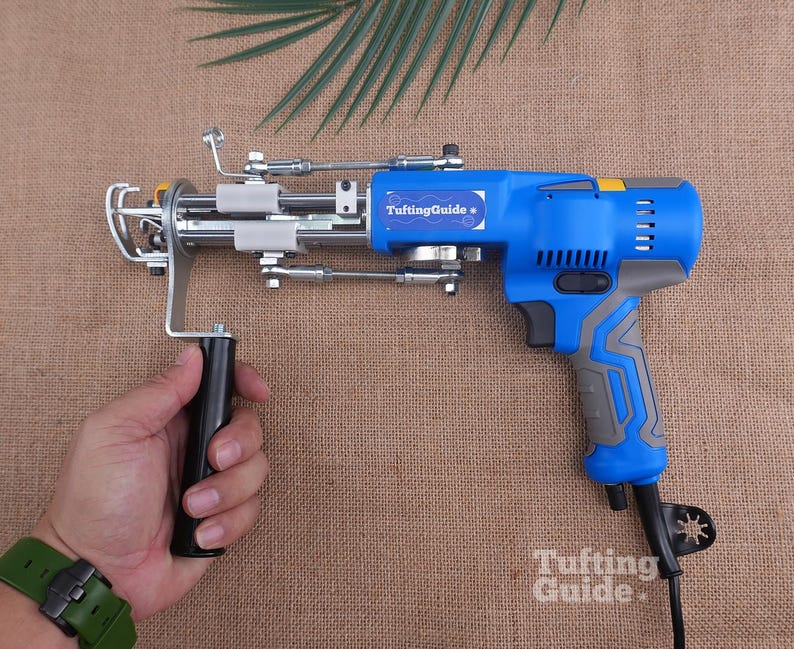May include: A blue and grey electric tufting gun with a metal needle mechanism and a black handle. The device features a blue label with the white text TuftingGuide and a small white star icon. The tool includes a black power cord and a trigger.