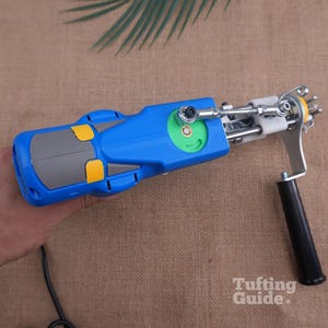 May include: A blue and grey electric tufting gun with yellow accents, a green circular gear component, a silver metal needle mechanism, and a black handle. The device features a white plastic part near the needle and a black power cord.