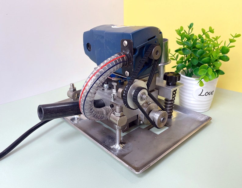 May include: A blue and silver machine with a black handle and a clear hose. The machine has a belt and pulley system. The machine is sitting on a silver metal base. The machine is on a white surface. There is a green plant in a white pot with the word "Love" on it in the background.