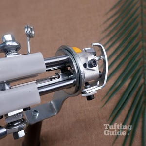 May include: A metal tufting gun mechanism featuring a silver-toned frame, two white plastic guide tubes, a yellow button, and exposed mechanical components including screws and a hook. The text Tufting Guide is visible in the bottom right corner.